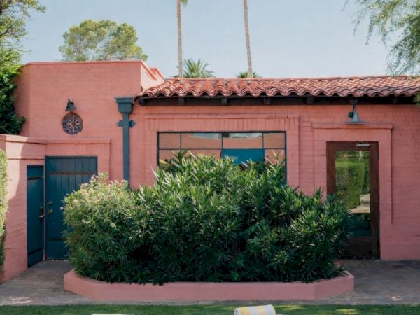 A pink stucco building with blue doors, palm trees, and neatly trimmed greenery; four yoga mats with towels laid on a grassy lawn.