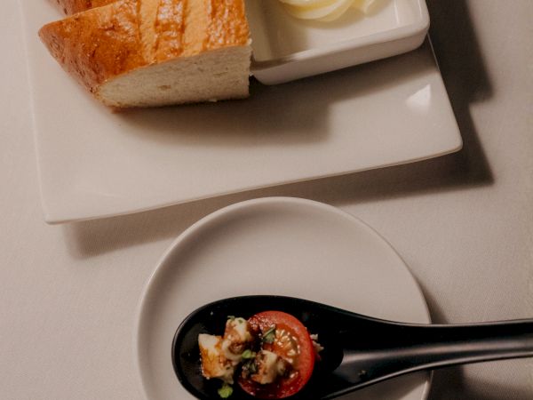 Slices of crusty bread with a small dish of butter, plus a spoon with a chunky tomato topping on a white plate and saucer.