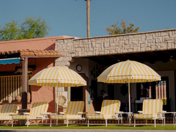 A sunny poolside scene with striped umbrellas, lounge chairs, and a tall palm tree behind a low stone building.