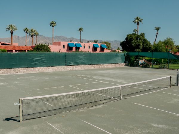 A deserted outdoor tennis court with a low net, faded lines, and a sunny backdrop of palm trees and colorful, low-rise buildings beyond a green wall.