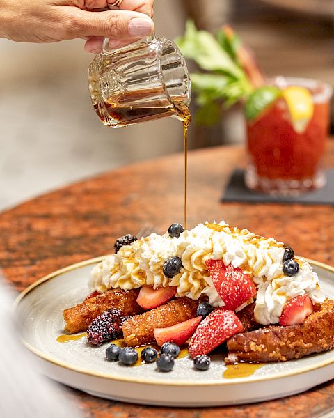 Golden syrup drizzled over a fruit-topped breakfast plate with berries, toast, and whipped cream, as a person pours syrup. End with period.