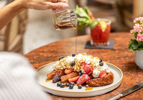 Golden syrup drizzled over a fruit-topped breakfast plate with berries, toast, and whipped cream, as a person pours syrup. End with period.
