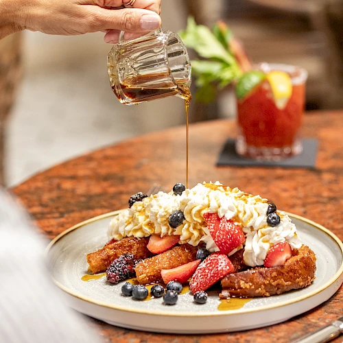 Golden syrup drizzled over a fruit-topped breakfast plate with berries, toast, and whipped cream, as a person pours syrup. End with period.