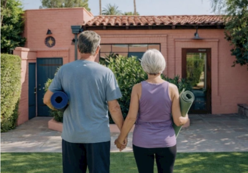 A couple holds hands with yoga mats, standing in front of a pink stucco house, ready for a sunny outdoor workout.
