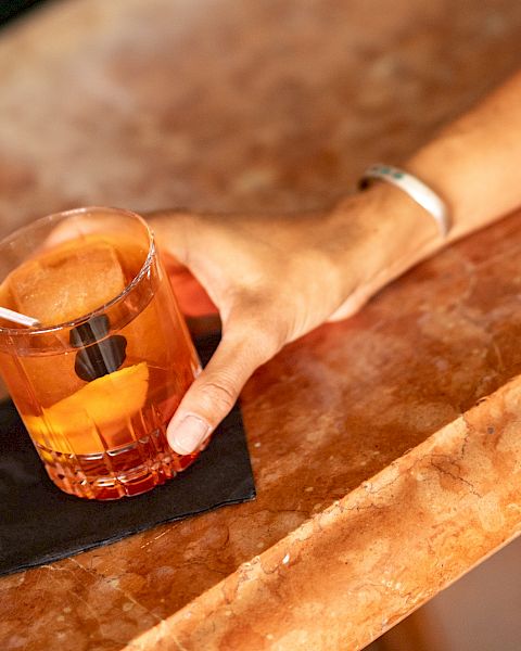 A hand rests on a bar counter with a short glass of amber liquid and a small black napkin beneath, suggesting a casual drink.