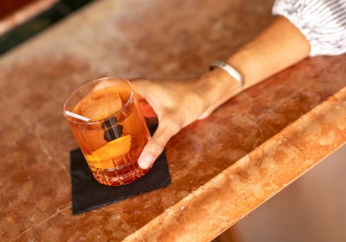 A hand rests on a bar counter with a short glass of amber liquid and a small black napkin beneath, suggesting a casual drink.