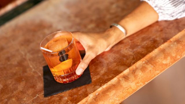 A hand rests on a bar counter with a short glass of amber liquid and a small black napkin beneath, suggesting a casual drink.