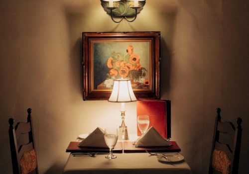 A cozy, dimly lit restaurant table for two with a lamp, framed art, and neatly folded napkins under warm ambiance.
