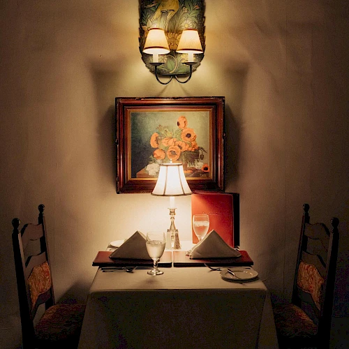 A cozy, dimly lit restaurant table for two with a lamp, framed art, and neatly folded napkins under warm ambiance.