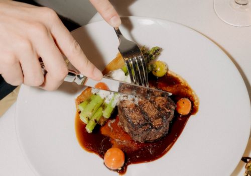 A person is cutting into a plated steak with sauce, accompanied by vegetables and a glass of wine on the side, dining elegant.