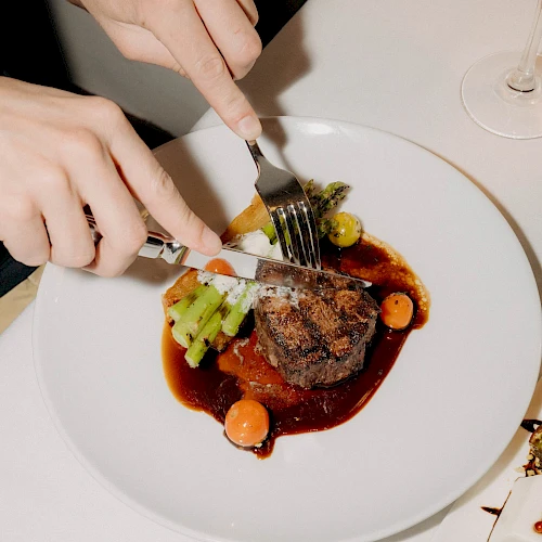 A person is cutting into a plated steak with sauce, accompanied by vegetables and a glass of wine on the side, dining elegant.