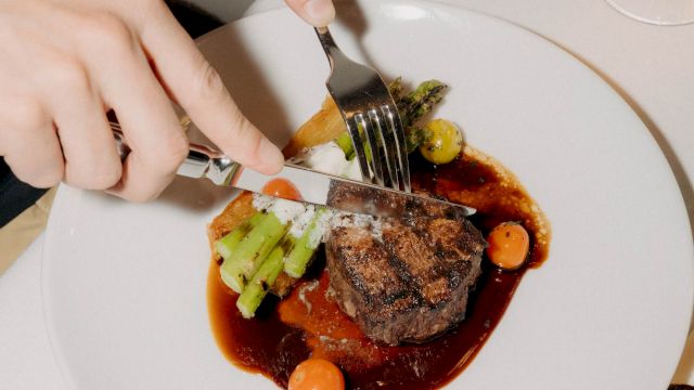 A person is cutting into a plated steak with sauce, accompanied by vegetables and a glass of wine on the side, dining elegant.