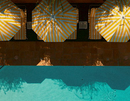 A sunny poolside scene with striped yellow-blue umbrellas, lounge chairs, and a blue water edge casting shadows over the pool.