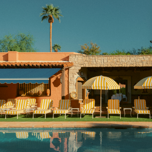 A sunny poolside scene with orange striped lounge chairs, yellow umbrellas, a stone wall building, and a clear blue sky, reflecting in the water.