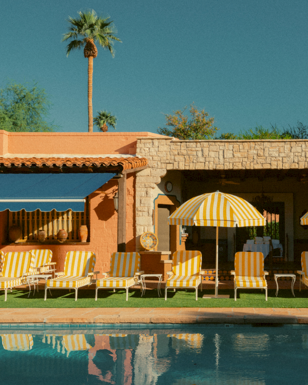 A sunny poolside scene with orange striped lounge chairs, yellow umbrellas, a stone wall building, and a clear blue sky, reflecting in the water.