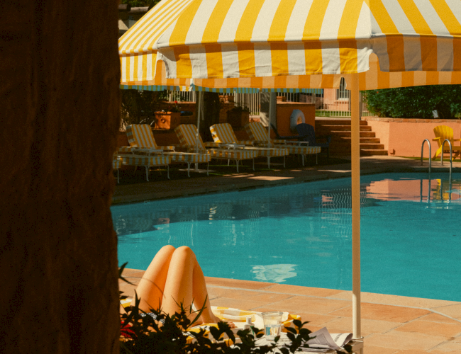 A person lounging by a pool under a yellow striped umbrella, sun chairs in the background, calm vacation vibe.
