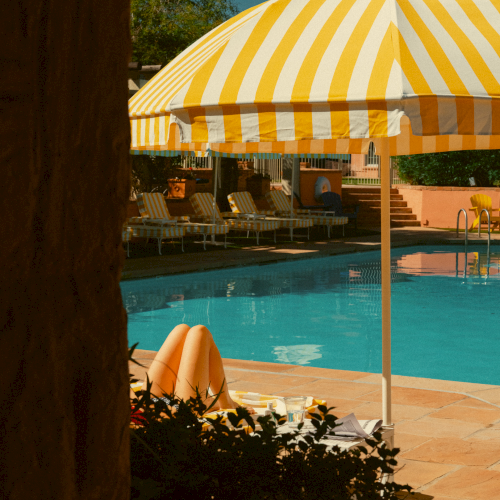 A sunny poolside scene with striped yellow umbrellas, lounge chairs, and clear blue water, partially shaded by a tree.