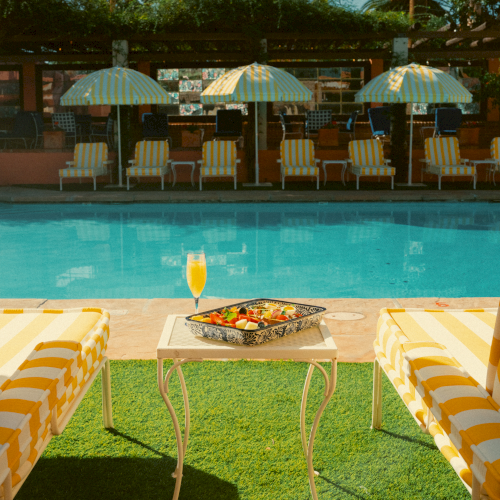 A sunny poolside scene with striped chairs, a small table holding a tray of food and a drink, and umbrellas in the background.