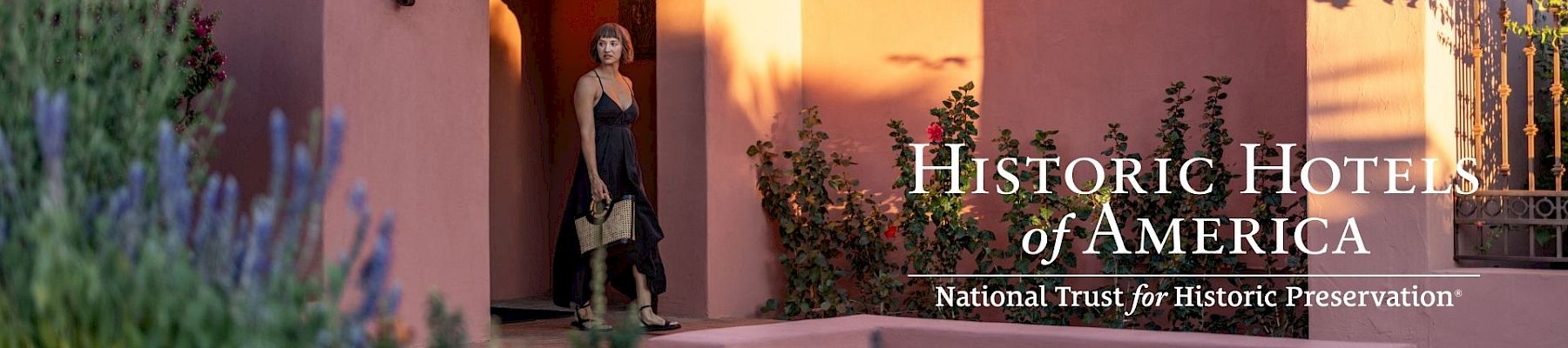 A woman stands at a shaded doorway of a peach-colored adobe building, framed by blooming plants, with &ldquo;Historic Hotels of America&rdquo; logo.
