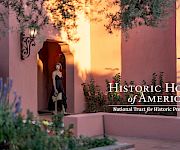 A woman stands at a shaded doorway of a peach-colored adobe building, framed by blooming plants, with “Historic Hotels of America” logo.
