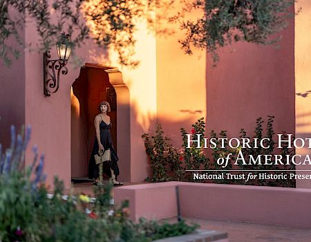 A woman stands at a shaded doorway of a peach-colored adobe building, framed by blooming plants, with &ldquo;Historic Hotels of America&rdquo; logo.