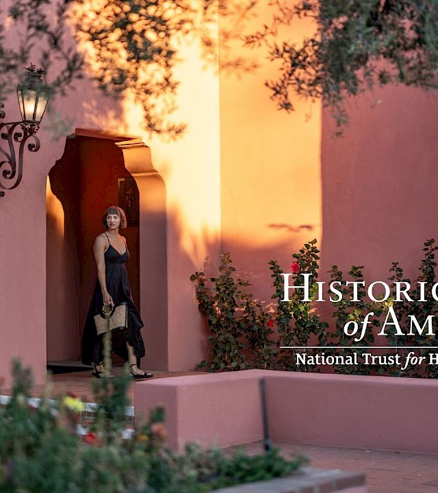 A woman stands at a shaded doorway of a peach-colored adobe building, framed by blooming plants, with “Historic Hotels of America” logo.