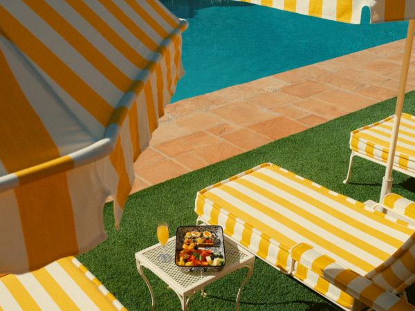A sunny poolside scene with yellow-and-white striped lounge chairs, a matching umbrella, and a small table holding snacks or fruit.