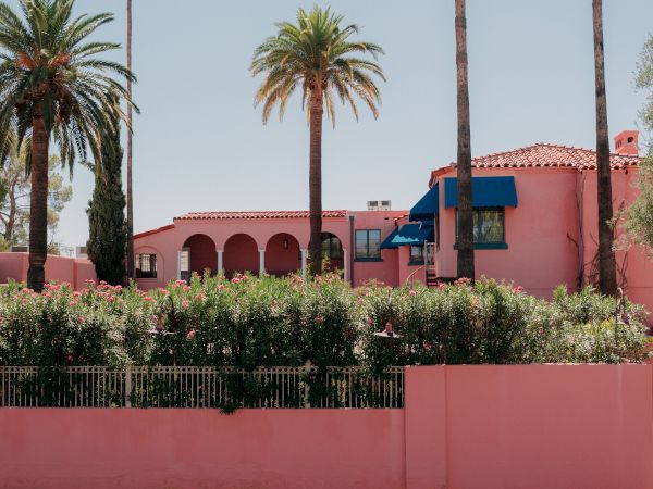 A pink Mediterranean-style house with arches, palm trees, a red tile roof, and a pink wall along a sidewalk.