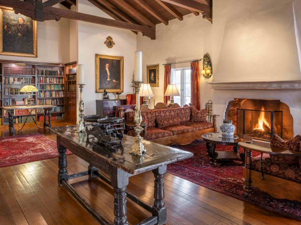 A cozy, classic living room with a stone fireplace, wooden beams, a large sofa and armchairs, a long coffee table, wood floor, and bookshelves.