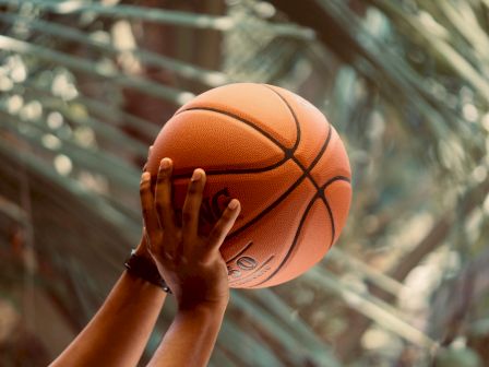 A person&rsquo;s hands grip an orange basketball on a court, with the background blurred and the ball in sharp focus.