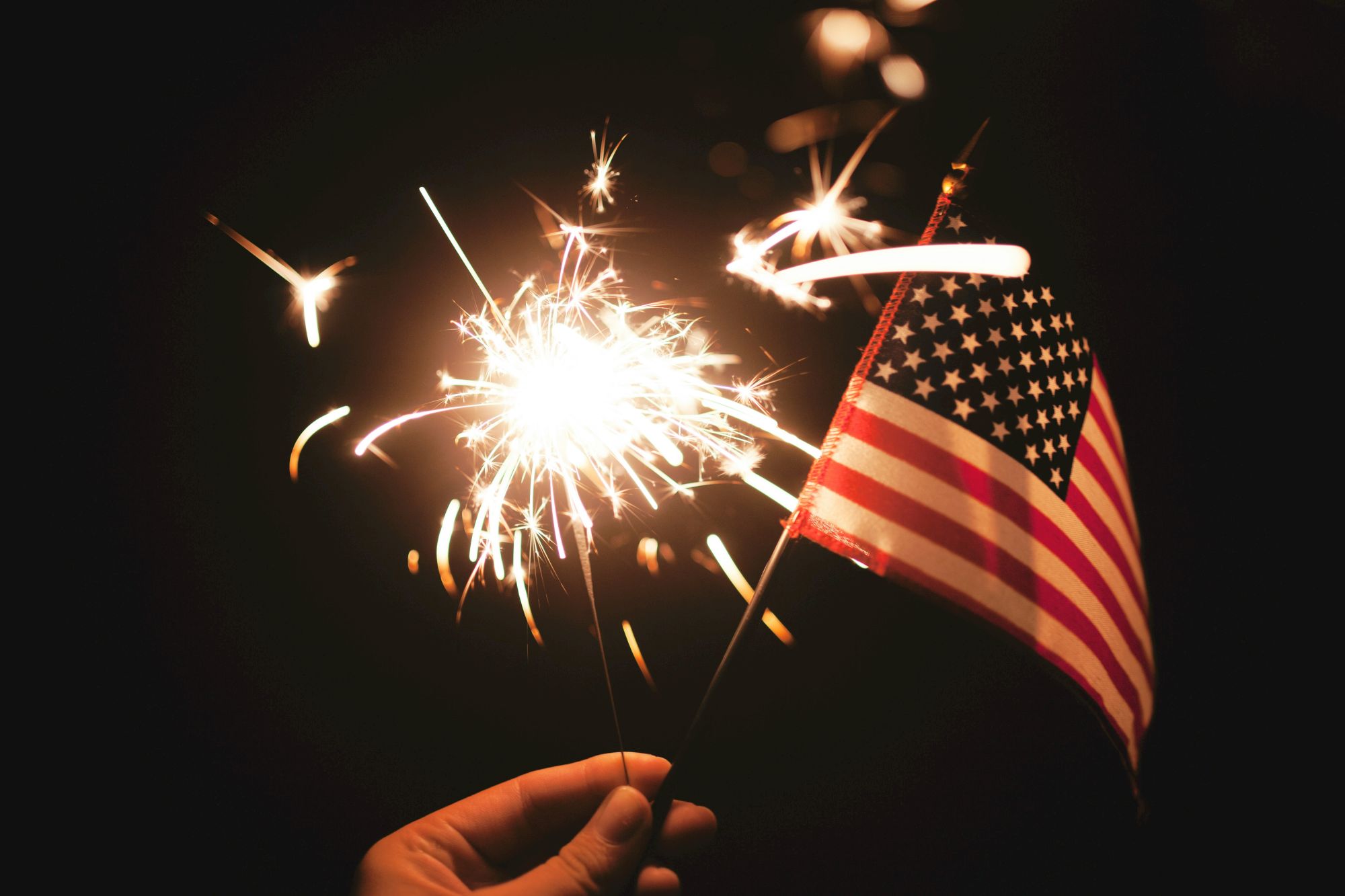 A hand holding a sparkler with bright sparks and a small American flag, fireworks-like glow against a dark background.