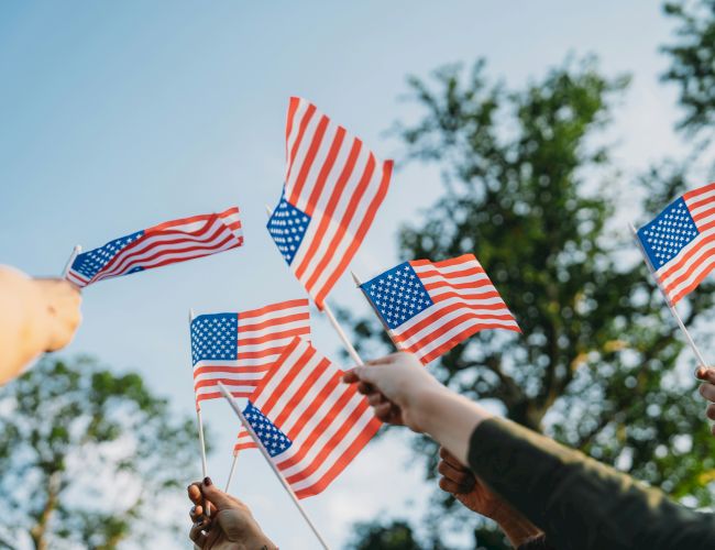 A group of people waving small American flags outdoors, flags fluttering against a blue sky with trees in the background.