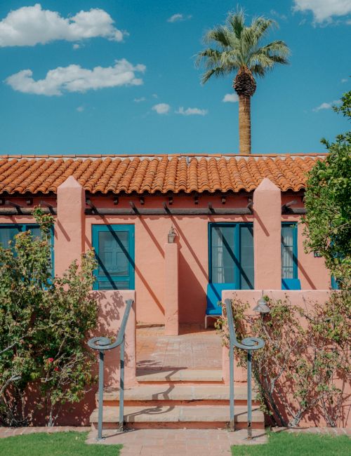 A pink single-story house with terracotta roof, blue doors/windows, a palm tree, and a sunny courtyard framed by pink pillars and greenery.