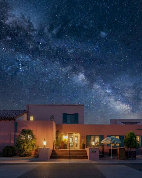 A desert-style house at night with warm lit windows under a starry sky and a visible Milky Way overhead.