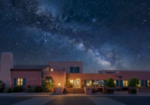 A desert-style house at night with warm lit windows under a starry sky and a visible Milky Way overhead.