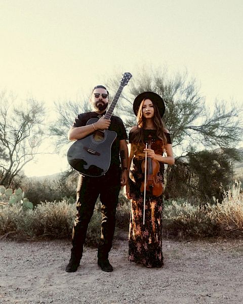 A man with a guitar and a woman with a violin stand in a desert landscape, dressed in casual, bohemian outfits, overcast sky.