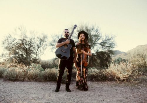 A man with a guitar and a woman with a violin stand in a desert landscape, dressed in casual, bohemian outfits, overcast sky.