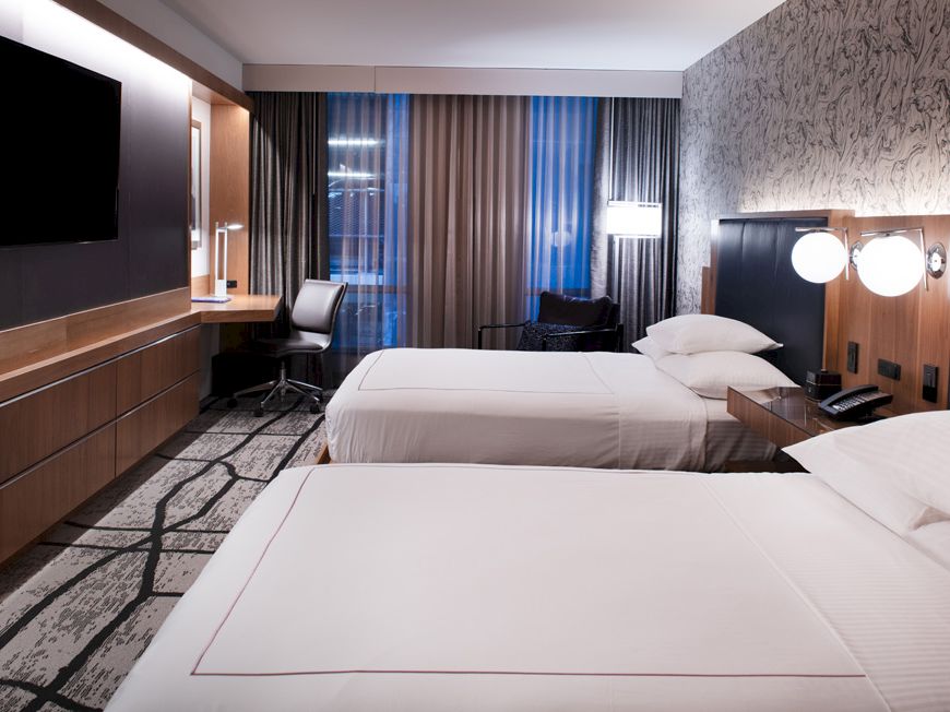 A modern hotel room with two neatly made beds, a wall TV, wood paneling, and floor-to-ceiling curtains by the windows.