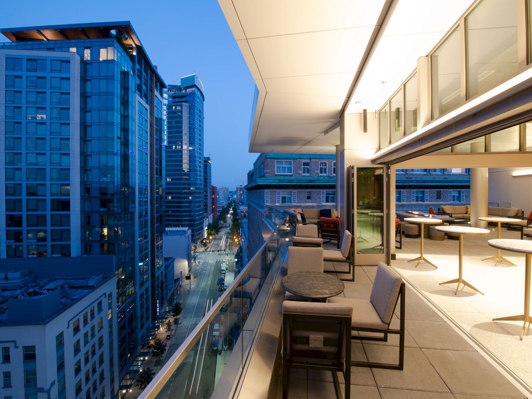A high-rise rooftop terrace with outdoor seating overlooks a city skyline and street at dusk.