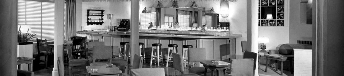 A vintage lounge and dining room with upholstered chairs, a tiled floor, and a long bar/counter under ornate trim, inside a house.