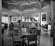 A vintage lounge and dining room with upholstered chairs, a tiled floor, and a long bar/counter under ornate trim, inside a house.