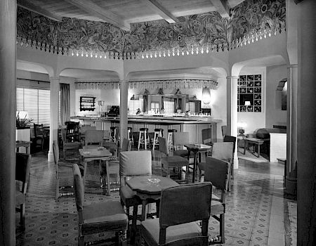 A vintage lounge and dining room with upholstered chairs, a tiled floor, and a long bar/counter under ornate trim, inside a house.