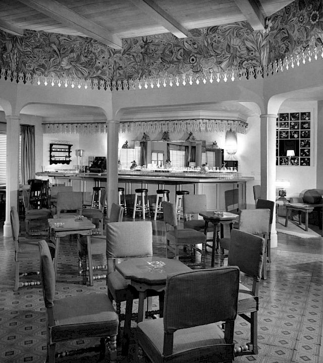 A vintage lounge and dining room with upholstered chairs, a tiled floor, and a long bar/counter under ornate trim, inside a house.