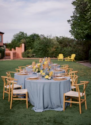 Outdoor event setup: round table with blue tablecloth, glassware, plates, and a colorful flower centerpiece on a lawn.