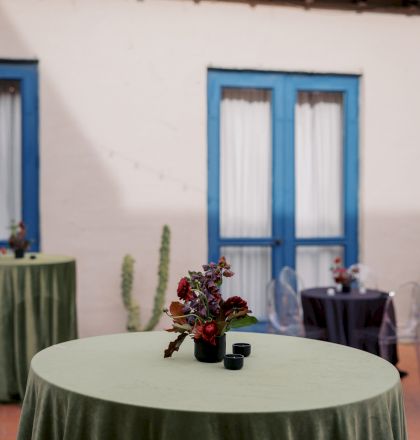 A round table with a green cloth centerpiece of red flowers sits in front of a blue-framed window at an event.