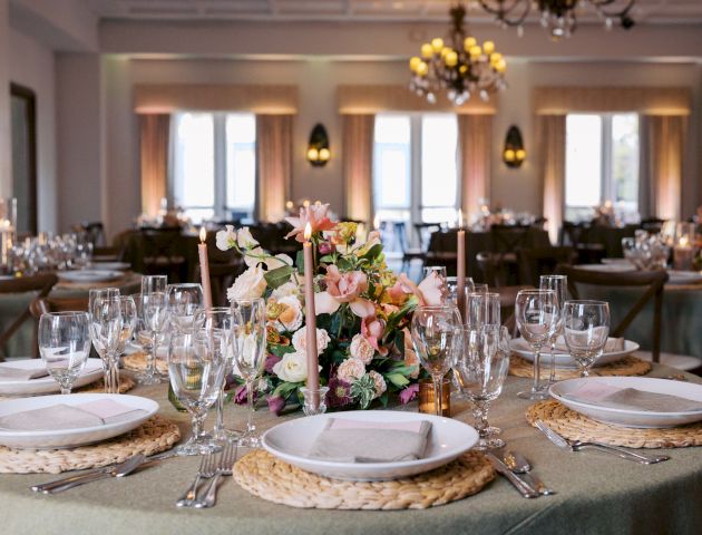 A decorated banquet table with floral centerpieces, lit candles, crystal glasses, and napkins in an elegant hall.