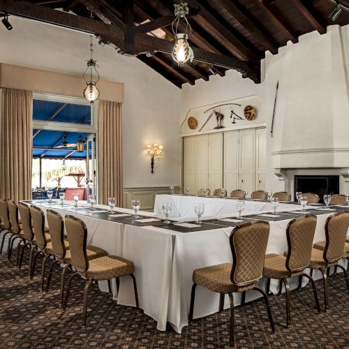 A formal banquet hall with a long white table, set glasses, cushioned chairs, chandeliers, and a fireplace on the wall.