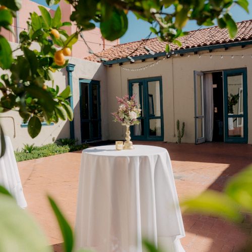An outdoor patio wedding setup with a white-clothed round table, flower centerpiece, string lights, and blue-framed doors and windows.