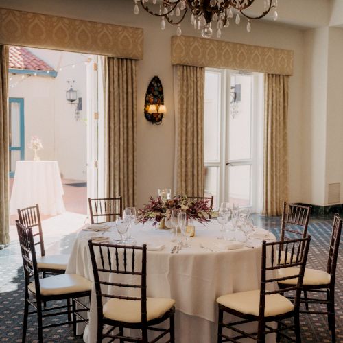 A banquet room set for an elegant event: round table with white linens, candles and flowers, under a sparkling chandelier and gold curtains.