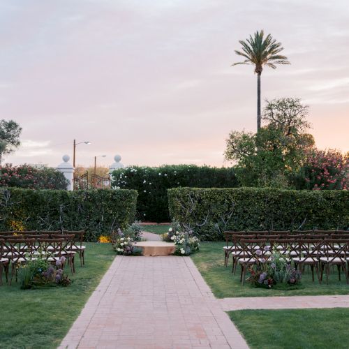 An outdoor garden wedding setup with rows of chairs, a flower-lined aisle, and a tall palm tree against a pastel sky.